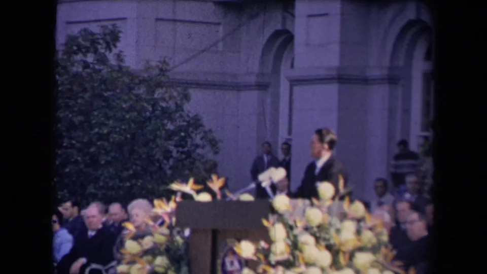 A side view shows the speaker at the decorated podium, with microphones, floral arrangements, seated attendees, and arched architecture in the background.