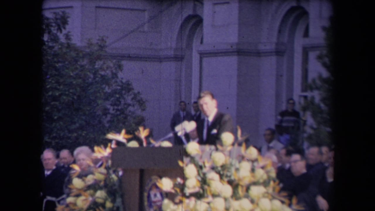 Reagan faces the audience from a flower-adorned podium, with microphones and formal attendees visible around him.