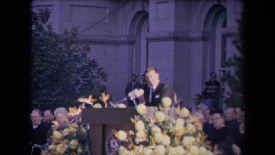 Reagan faces the audience from a flower-adorned podium, with microphones and formal attendees visible around him.