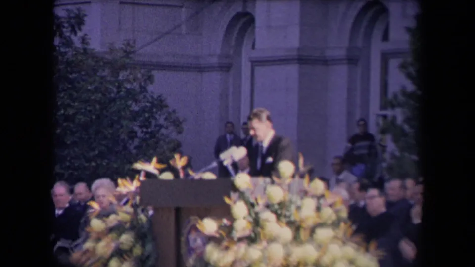 Reagan appears mid-speech beside clustered microphones, framed by yellow flowers and a seated formal audience.