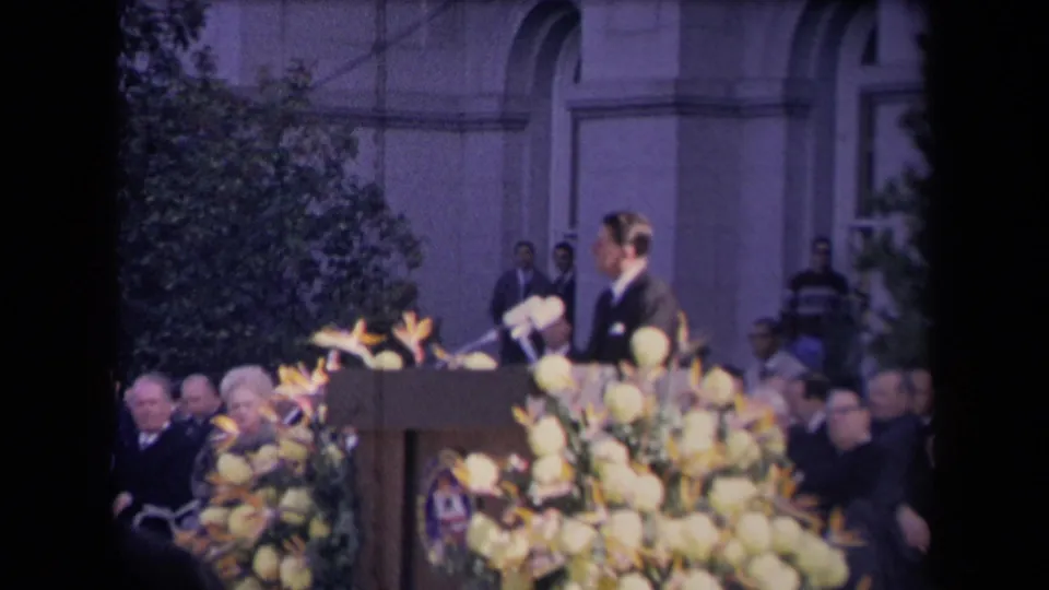 Reagan turns at the podium during the ceremony, with floral decorations, microphones, and seated attendees visible.