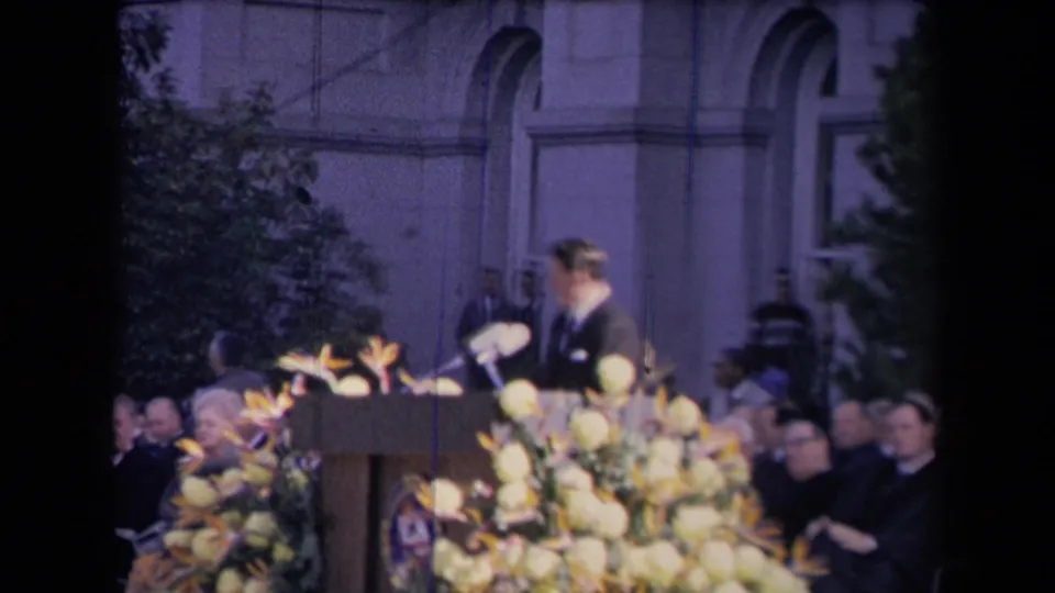 The camera catches the speaker in motion at the flower-lined podium, with formal attendees seated across the background.