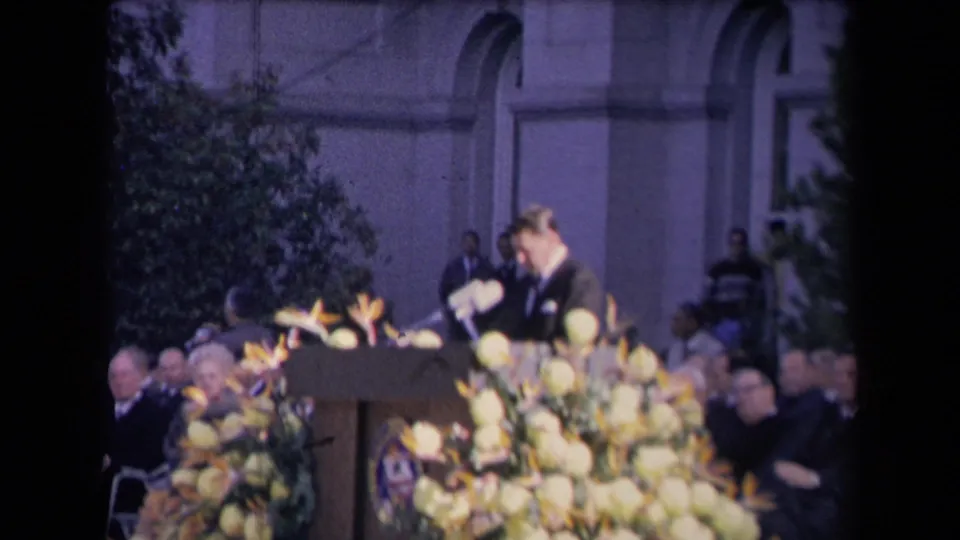 Ronald Reagan appears to look down toward notes or the podium, with microphones, flowers and formal guests visible around him.