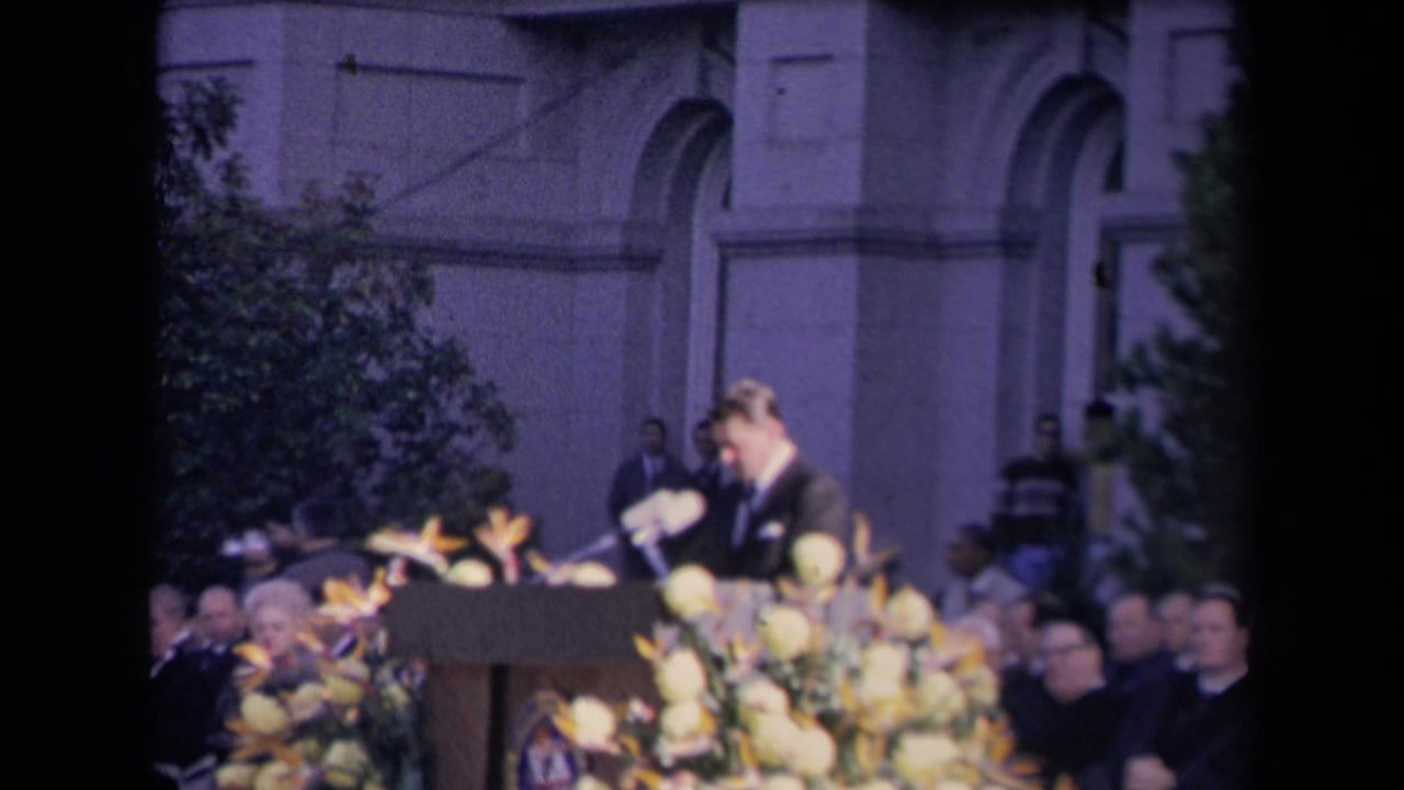 Suited speaker at a flower-covered podium before a seated audience and classical building facade.