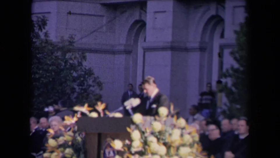A suited speaker appears at a podium with microphones and yellow flowers during an outdoor 1960s civic ceremony in Sacramento, per clip metadata.