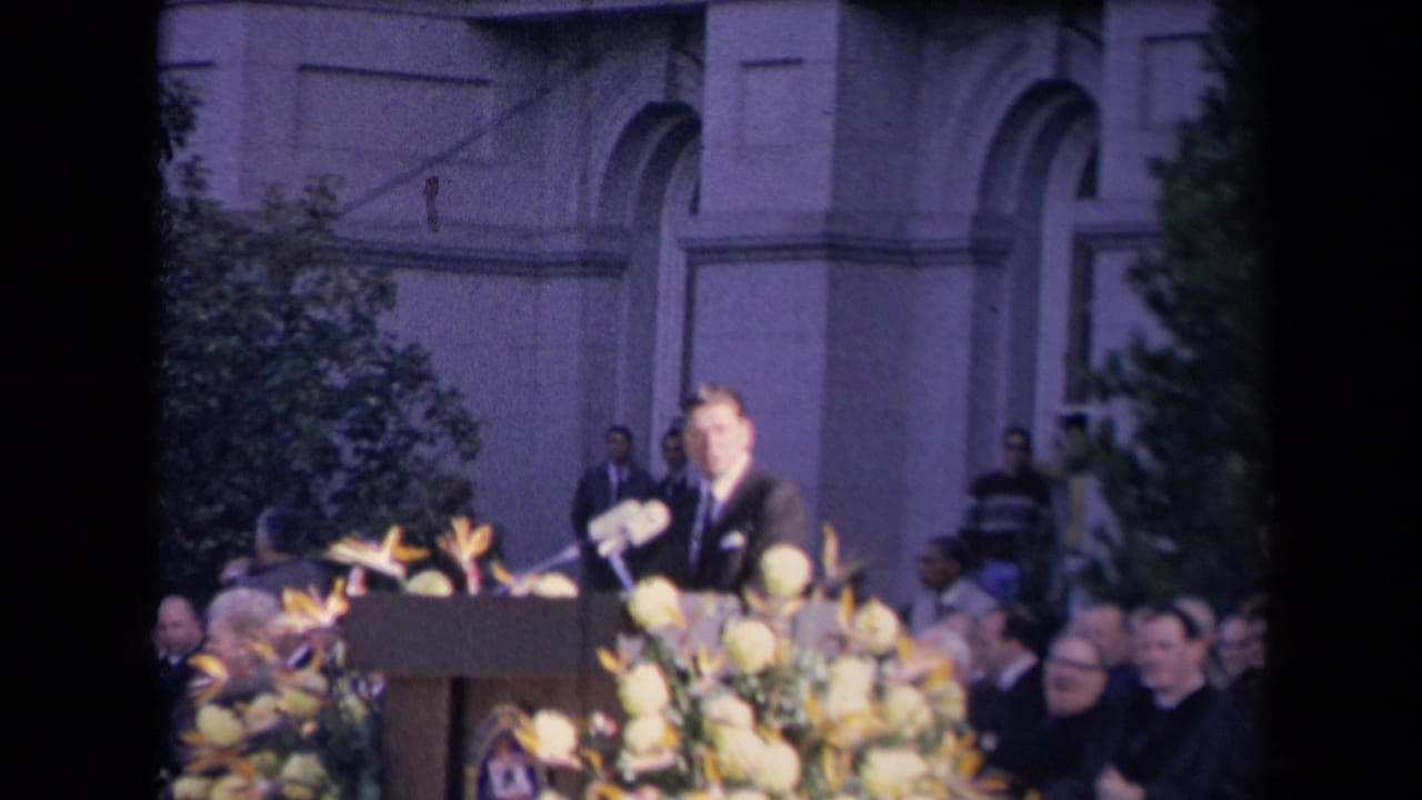 The speaker stands at the decorated podium as attendees watch from an outdoor stage area in the Reagan inauguration footage.