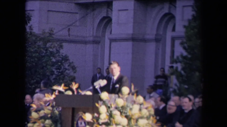 The speaker stands at the decorated podium as attendees watch from an outdoor stage area in the Reagan inauguration footage.