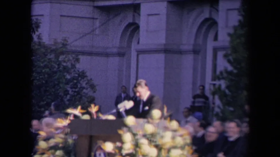 A soft 8mm frame shows the speaker leaning toward notes or the microphone, with flowers and audience members around the stage.
