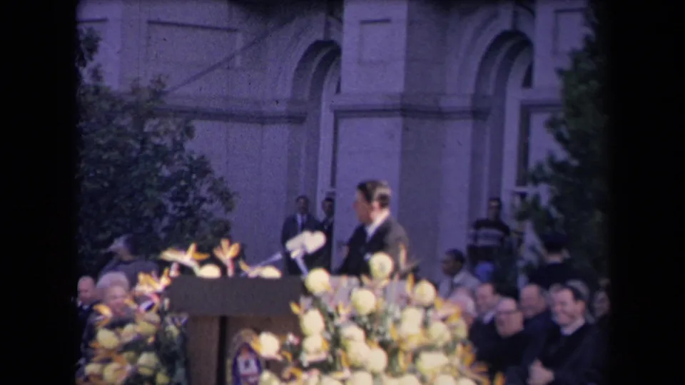 The speaker is seen in profile at the flower-covered podium, with formal attendees and the building facade behind him.
