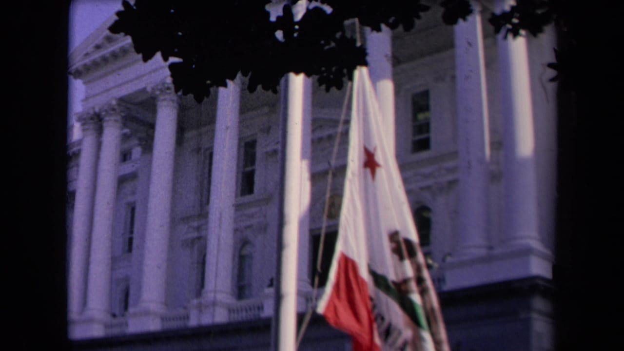 California state flag rising in front of a white-columned civic building under dark tree leaves.