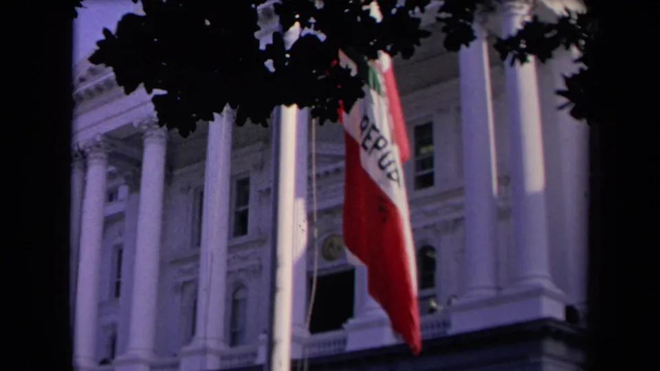A closer 8mm view catches the California flag crossing the frame, with columns, windows, and tree shadows in the background.