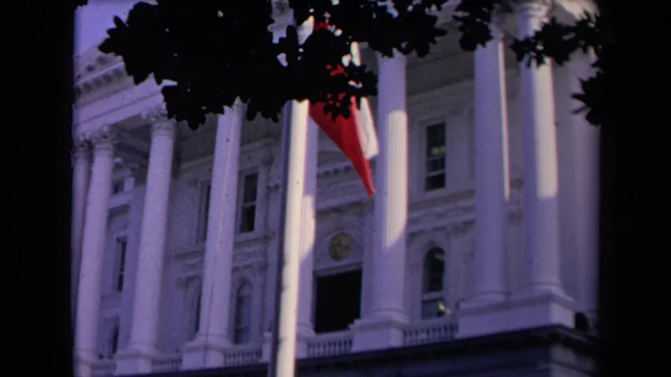 The flag is mostly turned away, leaving its red lower section visible against the columned building and dark overhead leaves.