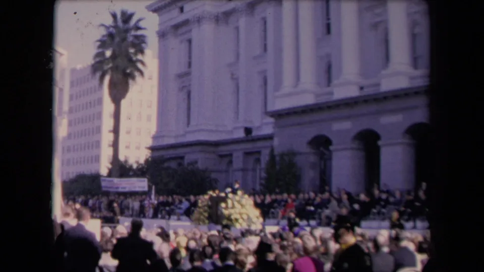 Handheld crowd view outside a formal civic-looking building, with attendees facing a stage or speaker area.
