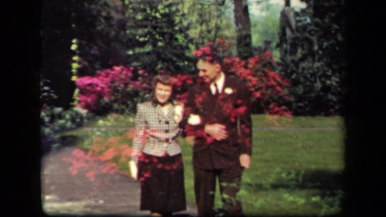 Adult couple walking arm-in-arm on a garden path with red flowers behind them.