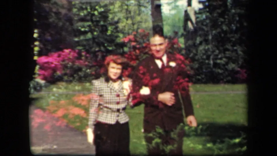 The couple continues toward the camera in the New York garden, with the man in a dark suit and the woman in a checked jacket.