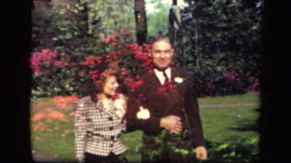 Close 1941 garden frame of the couple arm-in-arm, with the woman turned toward the man and red flowering shrubs behind them.