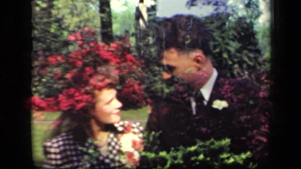 The couple pauses in the garden, looking toward each other among shrubs and bright red flowers in the 1941 New York clip.