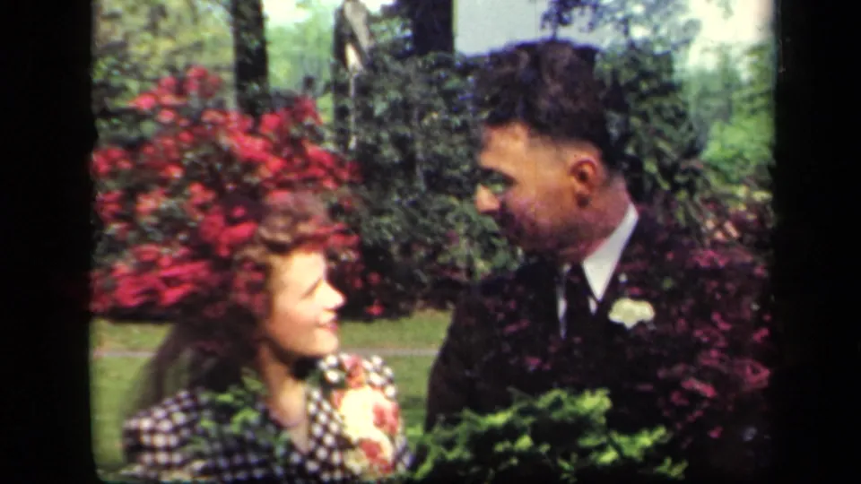 A closer home-movie frame shows the woman and suited man in conversation, with red flowers and greenery behind them.