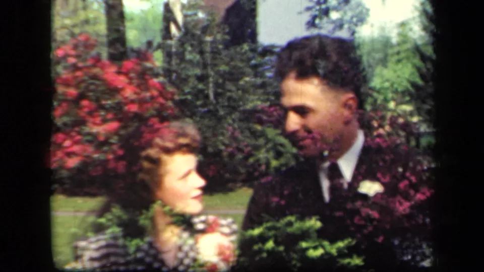 The suited man smiles toward the woman as they stand beside dense greenery and red flowering shrubs in the supplied New York footage.