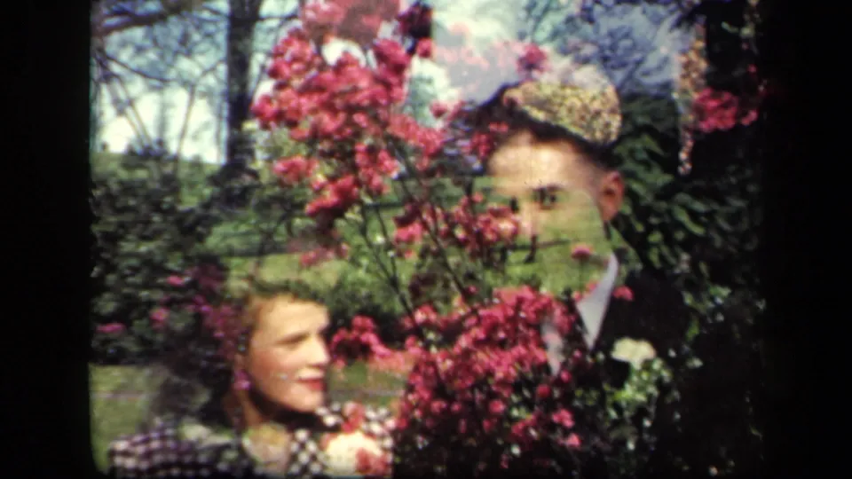 In 1941 New York home-movie footage, a woman in a checked dress and a man in a dark suit pose among pink flowers.