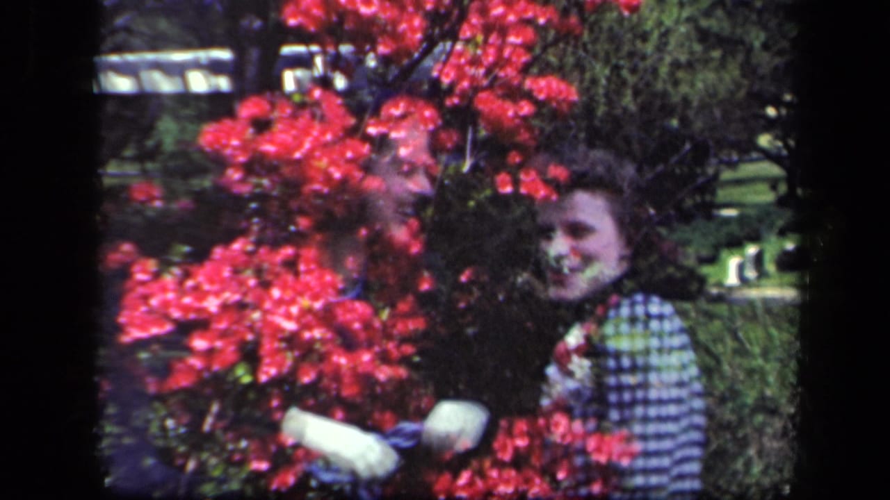 1941 New York home-movie frame with a smiling woman in a checkered dress partly overlaid with red garden flowers.