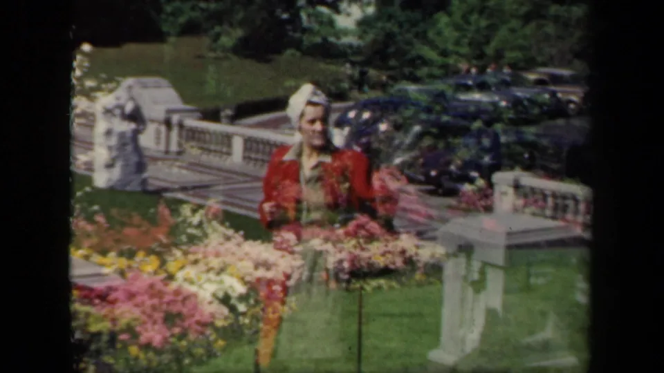 1941 New York 8mm frame showing a woman in a red dress and white hat posing by flower beds on formal grounds.