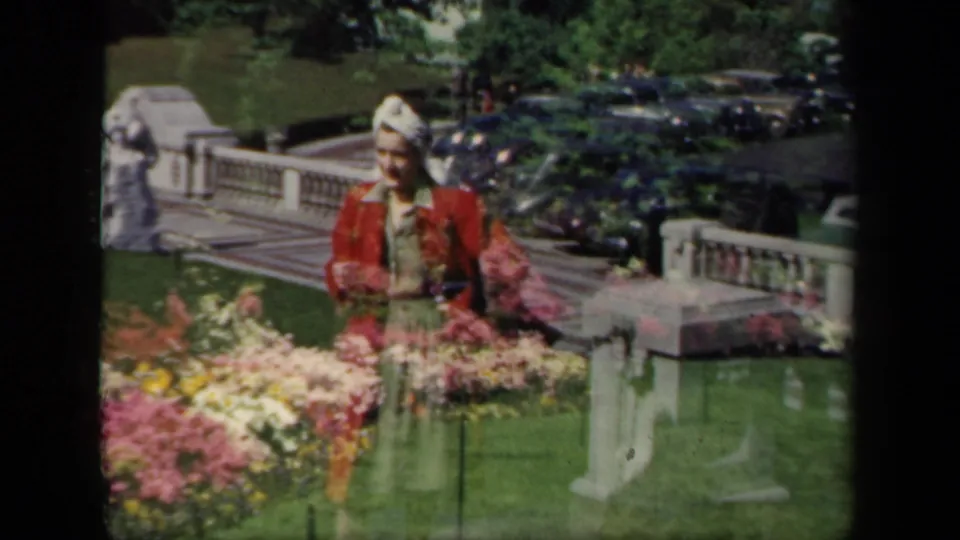 1941 New York home-movie frame of a woman in red standing beside flowers, with formal railing, lawn, and parked cars behind.