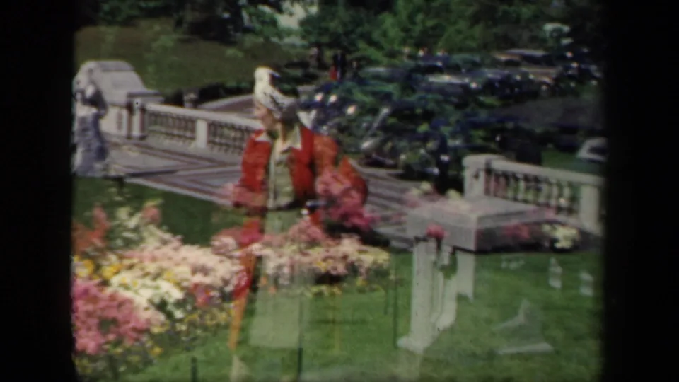 1941 New York home-movie frame showing a woman in a red jacket and white hat among colorful garden beds with stone railings and cars behind her.