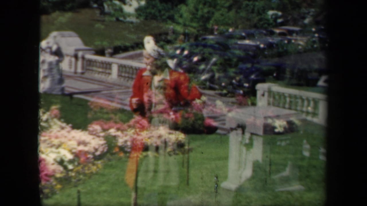 1941 New York garden scene with a woman in a red jacket and white hat standing near floral displays, stone balustrade, and parked cars.