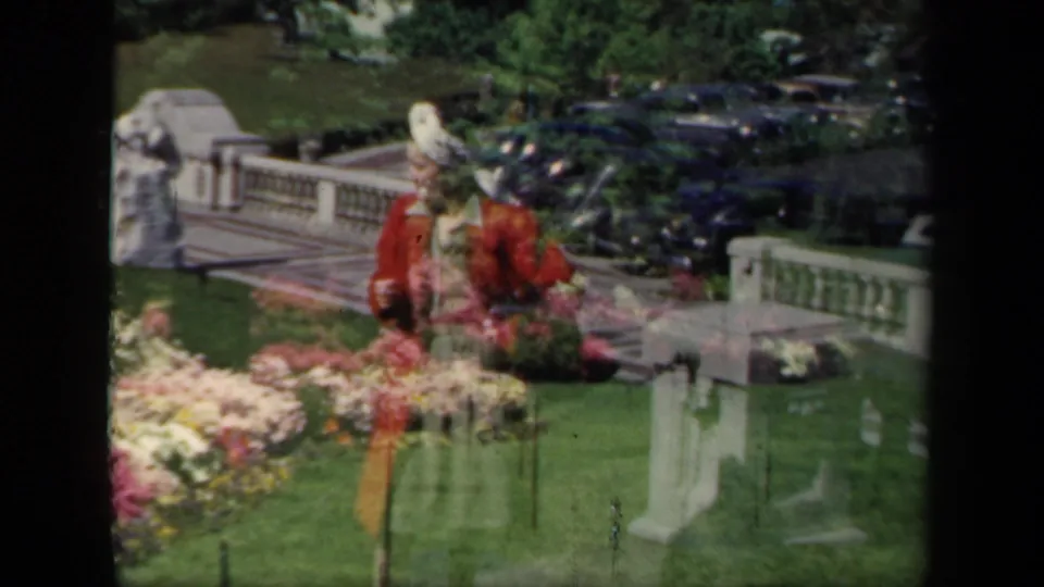 1941 New York garden scene with a woman in a red jacket and white hat standing near floral displays, stone balustrade, and parked cars.