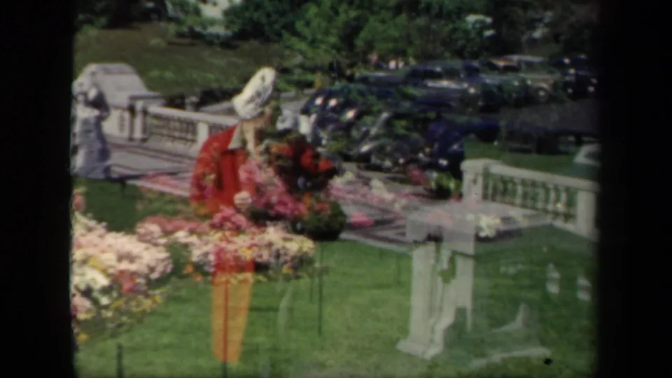 1941 New York home-movie frame of a woman bending or leaning near colorful flowers on formal garden grounds with parked cars behind.