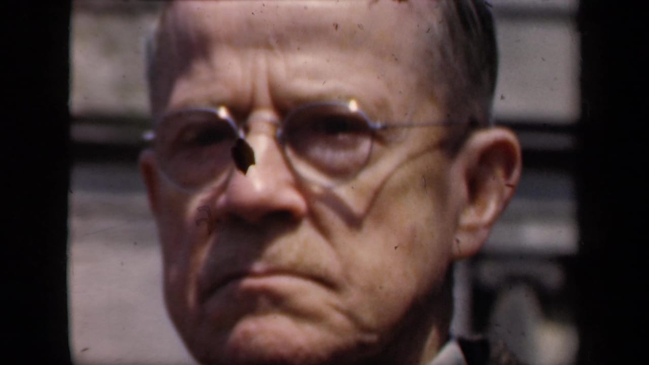1941 New York 8mm close-up of an older man in round glasses and a dark jacket, seen against a soft outdoor architectural background.
