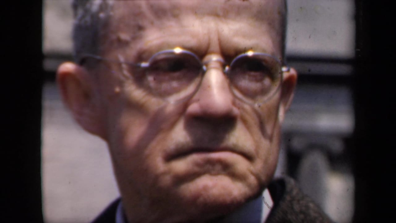 Older man with round glasses looking serious in close-up.