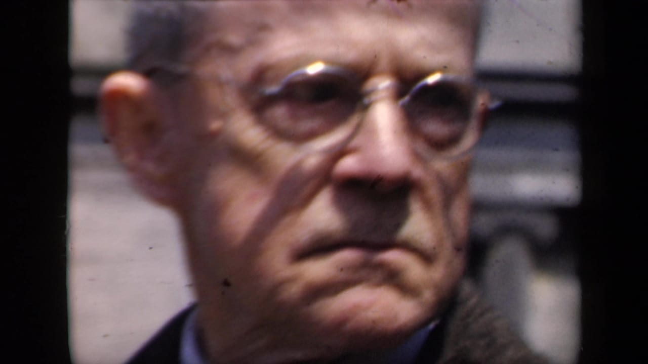 The same older man in round glasses turns slightly, with a stone building background behind him.