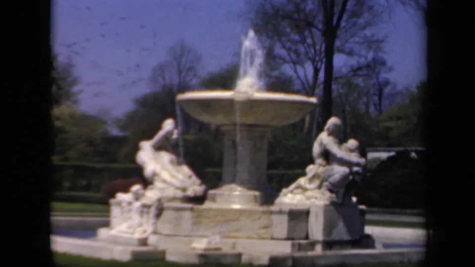 A sunlit public fountain with classical sculpted figures, trees, and blue sky in 1941 New York footage.