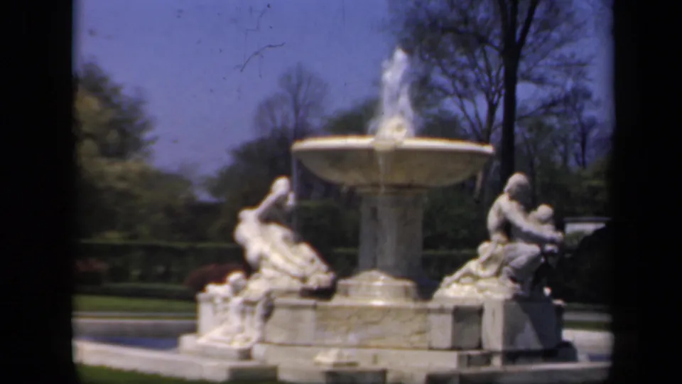 The fountain remains in view, with water spray, seated sculptures, trees, and a clear blue sky.