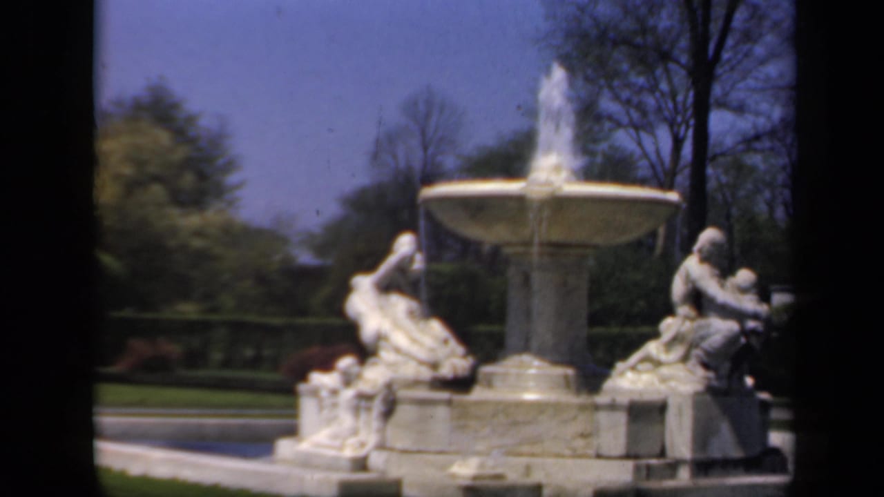 1941 8mm home-movie view of a stone garden fountain with water spray, reclining statues, lawn, trees, and blue sky in New York.