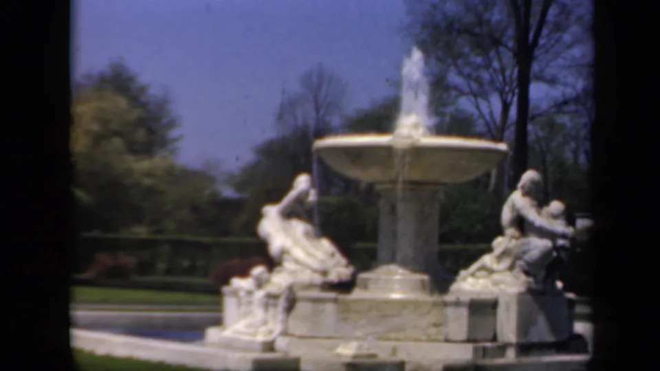 1941 8mm home-movie view of a stone garden fountain with water spray, reclining statues, lawn, trees, and blue sky in New York.