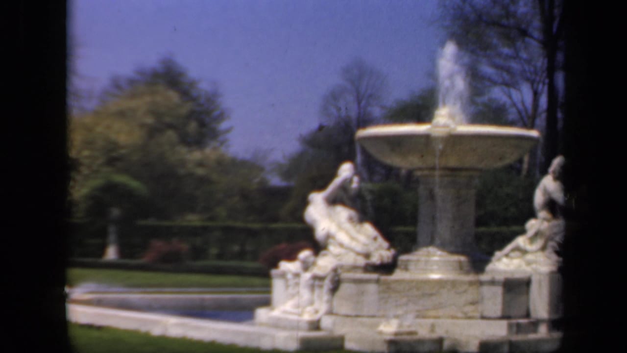 Stone fountain and reclining statues framed by lawn, trees, and blue sky.