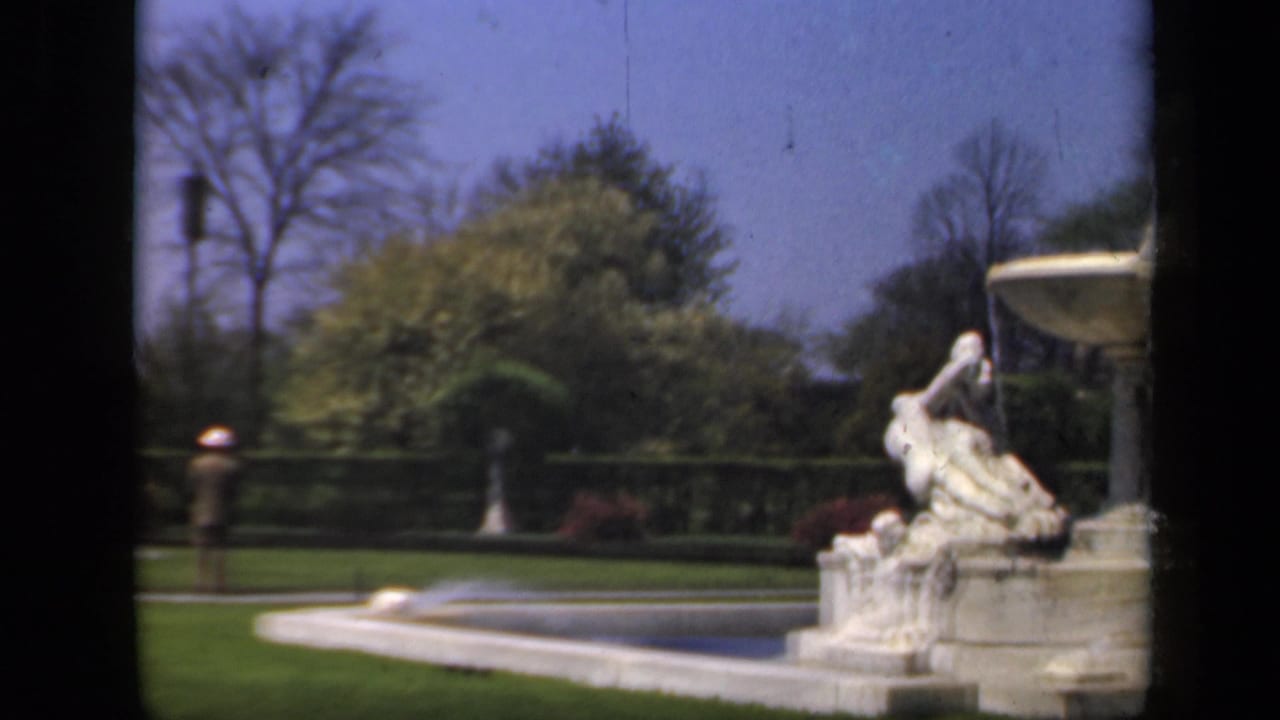 White ornamental fountain in a green garden with paths, trees, and a small distant figure.