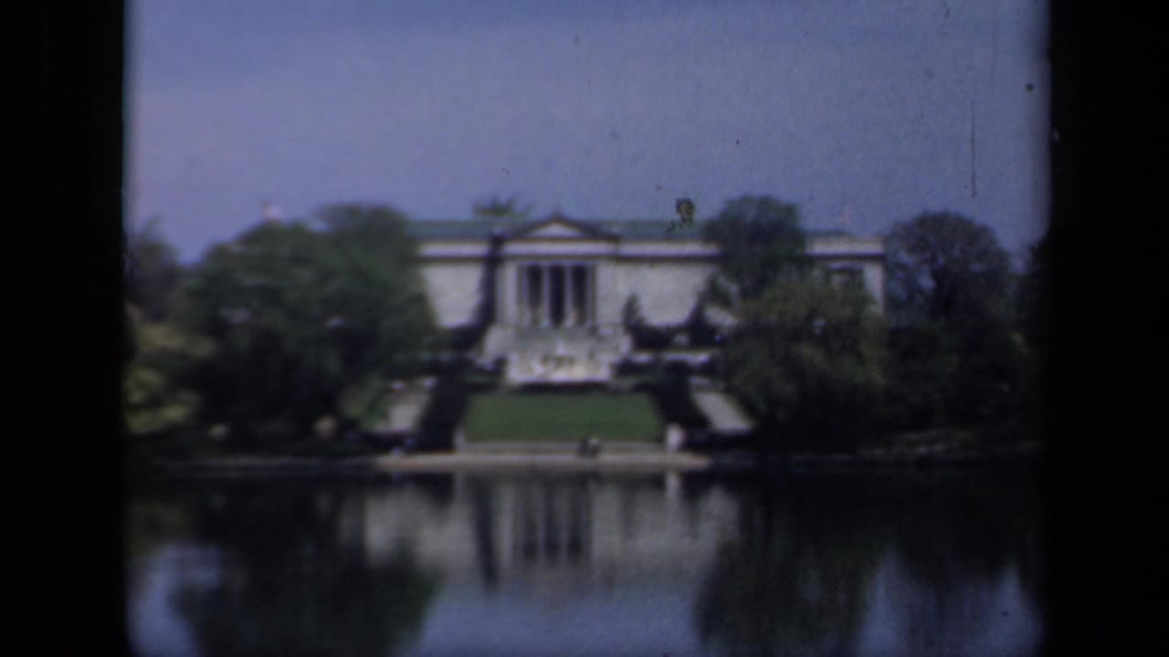 Columned building across a lake, framed by trees with a reflection in the water.