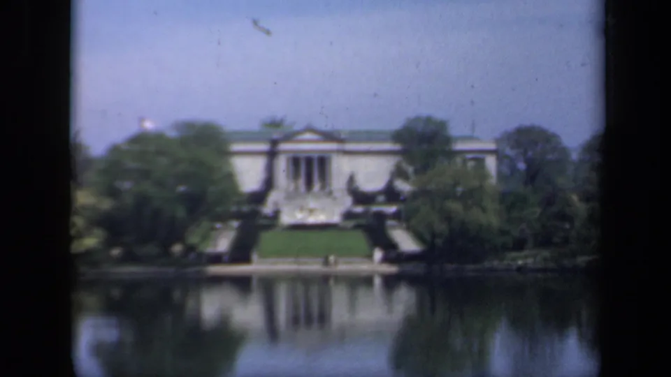 The home-movie frame shows a columned New York building, formal lawn, dense trees, and mirror-like lake reflection in 1941 footage.