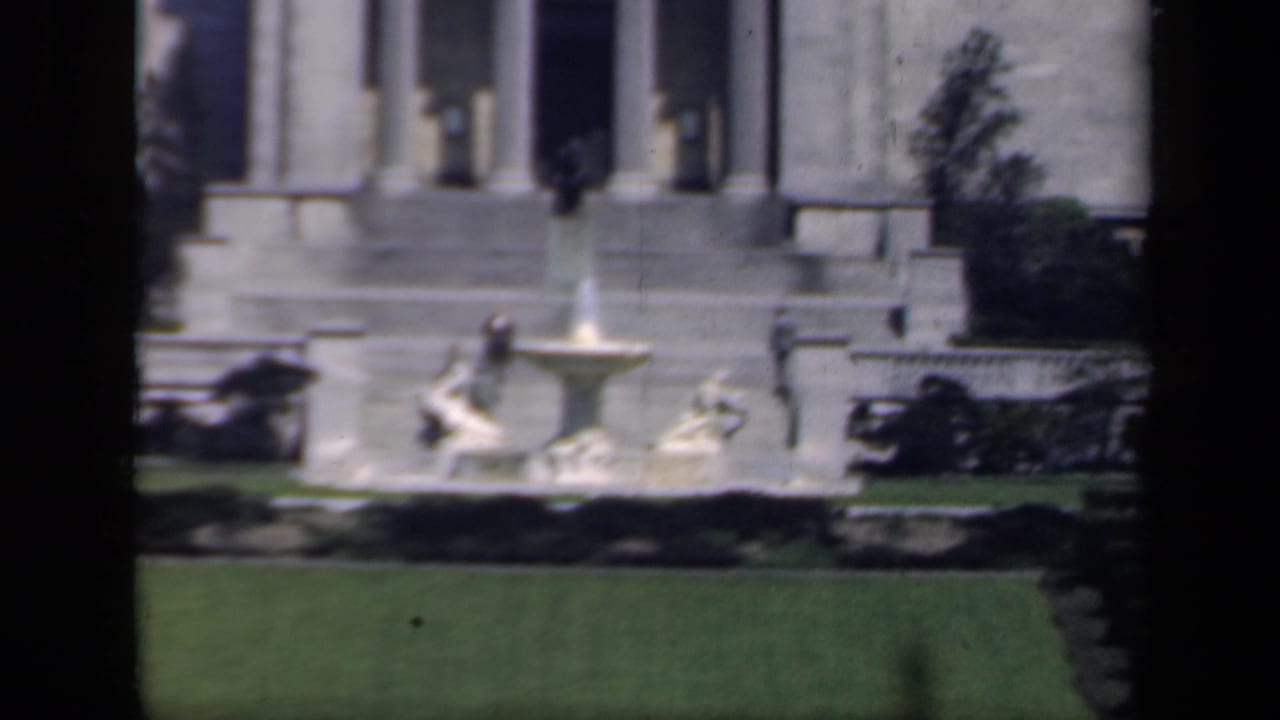 1941 New York frame with a formal fountain on the grounds of a classical stone building and small distant visitors nearby.