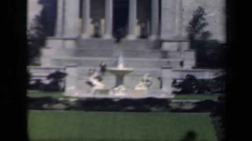 1941 New York home-movie frame of a fountain and classical building facade, with visible film gate borders and soft color.