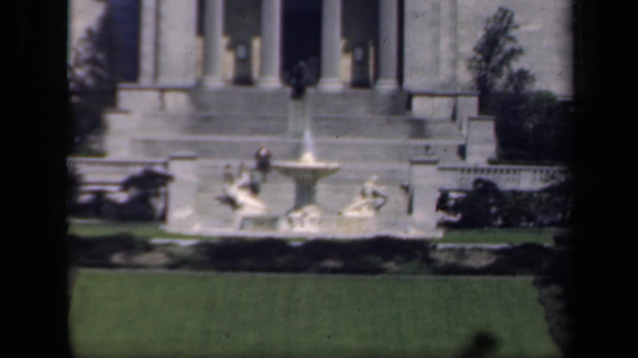 1941 New York home-movie frame showing a stone fountain, manicured lawn, shrubs, and a classical columned building.