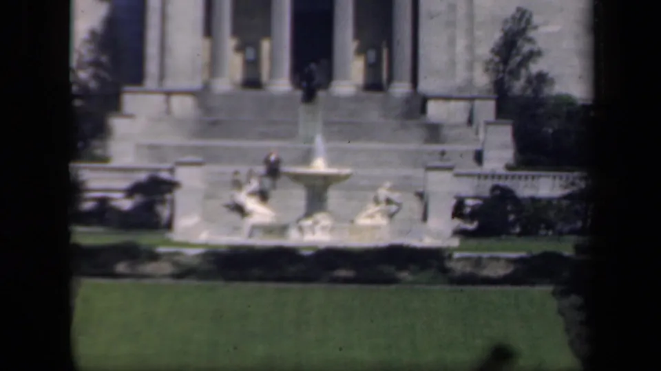 1941 New York home-movie frame showing a stone fountain, manicured lawn, shrubs, and a classical columned building.