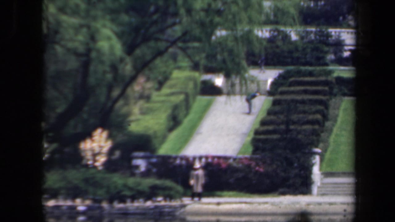 1941 New York garden view with clipped hedges, a pale walkway, stone steps, mature trees, and a distant walker.