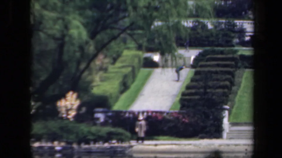 1941 New York garden view with clipped hedges, a pale walkway, stone steps, mature trees, and a distant walker.
