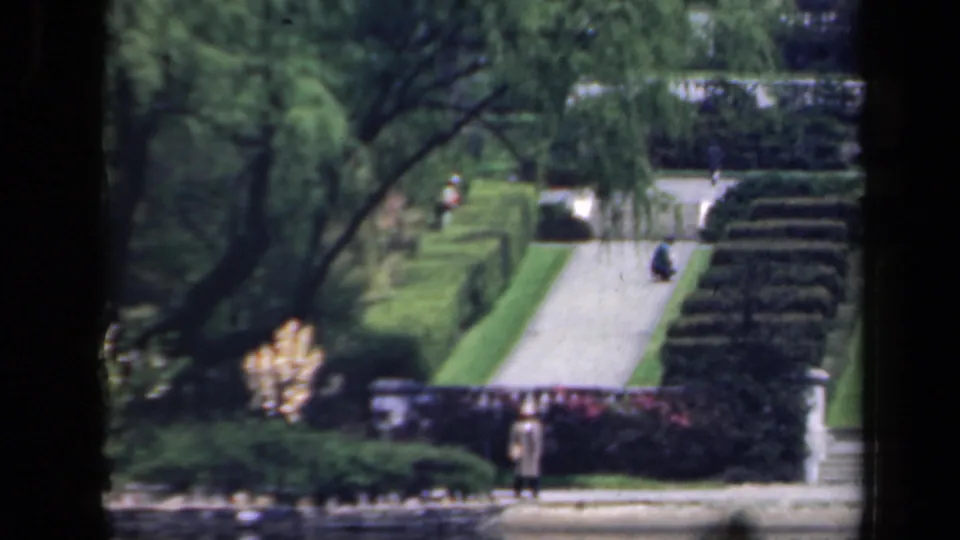 1941 New York garden footage showing a person near steps, with formal hedges, a bright walkway, and shaded trees behind.