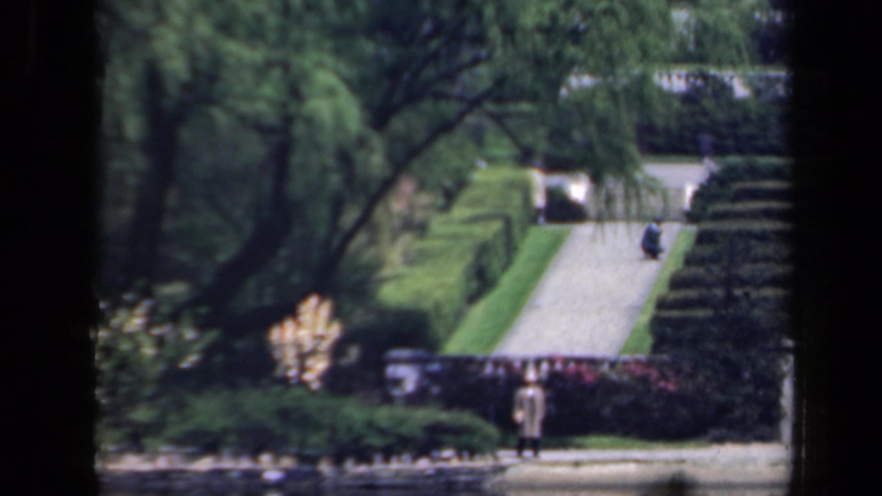 1941 New York home-movie frame showing a landscaped Japanese-style garden with large trees, clipped hedges, a pale path, and a distant visitor.
